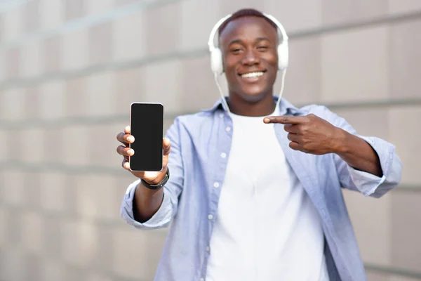 Красивий чорний чоловік в гарнітурі вказує на мобільний телефон з порожнім екраном на відкритому повітрі, макет для мобільного додатку або дизайну веб-сайту — стокове фото