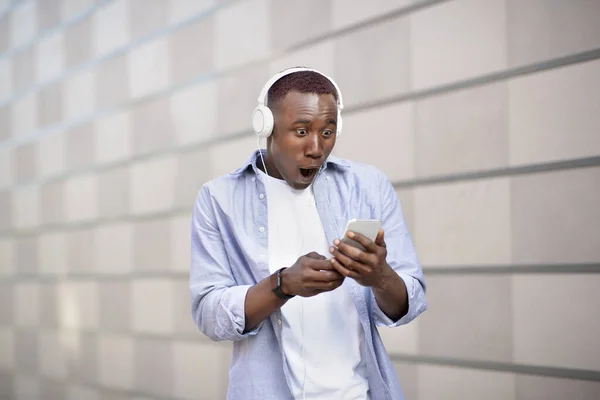 Chockad afroamerikansk man i headset stirrar på smartphone-skärmen på city street — Stockfoto
