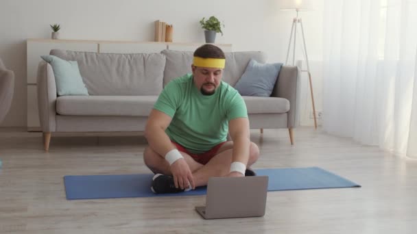 Hombre con sobrepeso sentado en posición de loto contra el portátil y meditando, practicando ejercicio respiratorio en el suelo en casa — Vídeos de Stock