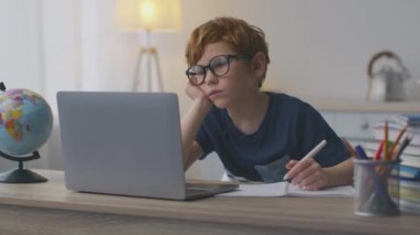 Sıkıcı online eğitim. Yorgun kızıl saçlı çocuk evde video dersi ya da webinar izliyor, dizüstü bilgisayara bakıyor ve yorgun hissediyor.