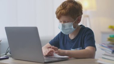 Koruyucu tıbbi maske takan küçük kızıl okul çocuğu evde çalışıyor, iş yerinde dizüstü bilgisayarla çalışıyor.