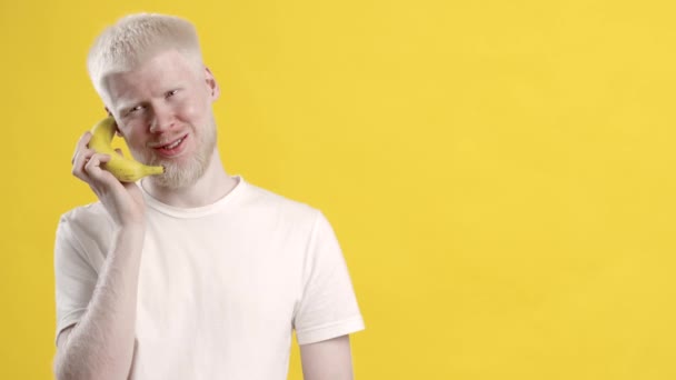 Позитивный альбинос разговаривает с банановым телефоном на жёлтом фоне — стоковое видео