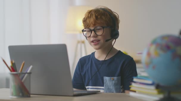Pieni punapää poika yllään kuulokkeet mikrofoni video chattailuun tutor, katselee kannettavaa tietokonetta ja puhuu opettaja — kuvapankkivideo