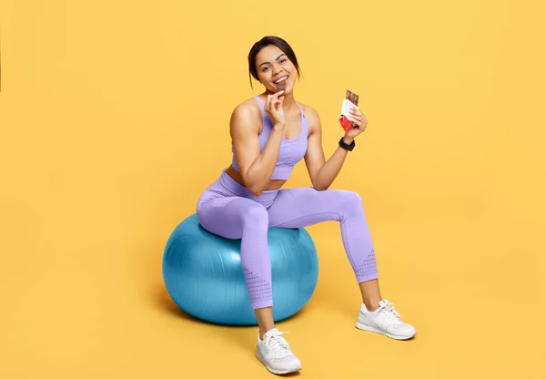 Mutlu Afro-Amerikalı kadın fitball 'da oturuyor ve çikolata yiyor, eğitimden sonra ara veriyor, sarı arka plan — Stok fotoğraf