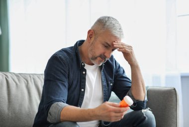 Yaşlı adamın başı ağrıyor, elinde haplarla kavanoz tutuyor.