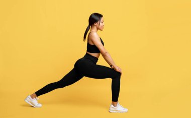 Spor kıyafetli Afrikalı Amerikalı kadın antrenmandan önce bacaklarını esnetiyor, sarı arka planda spor eğitimine hazırlanıyor.