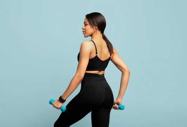 건강에 대한 개념. 젊은 아프리카계 미국 여성 이 두 개의 덤벨을 가지고 운동을 하고 푸른 배경 위로 돌아눕는 모습 — 스톡 사진