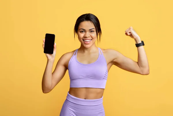 Aplicação de fitness. Mulher negra feliz segurando smartphone com tela em branco e mostrando músculos do braço, fundo amarelo — Fotografia de Stock