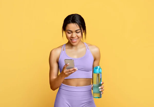 Gadget moderni per l'allenamento. Felice donna nera in possesso di bottiglia con acqua e utilizzando smartphone, sfondo giallo — Foto Stock