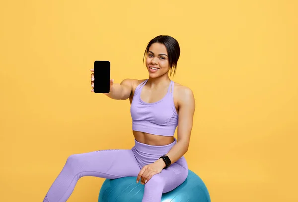 App för träning. Glad svart dam som håller mobiltelefon med svart skärm, sitter på fitball över gul bakgrund — Stockfoto