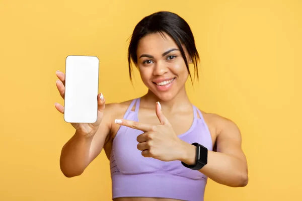 Sports application. Excited black lady holding and showing smartphone, indicating at black empty screen for mockup