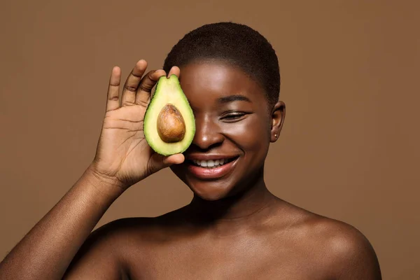 Aantrekkelijke naakt zwart vrouw met schone huid Covering oog met avocado helft — Stockfoto