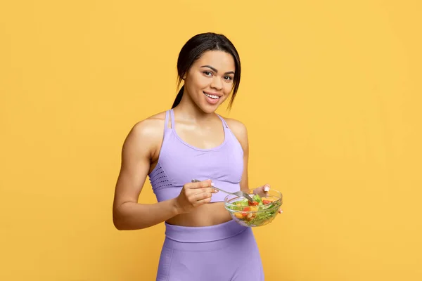 Healthy food concept. Fit african american woman eating fresh vegetable salad, smiling to camera over yellow background — Stock Photo, Image