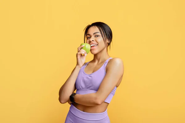 Zwarte sportieve dame met goed fit lichaam bijten appel, het eten van vers fruit na de training, staande ober gele achtergrond — Stockfoto