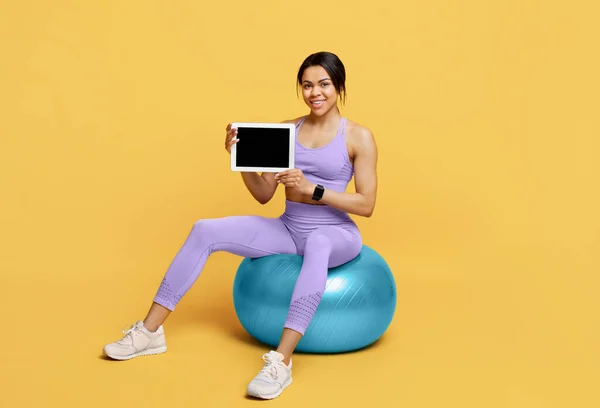 Deportiva mujer afroamericana mostrando tableta con pantalla vacía en fitball sobre fondo amarillo —  Fotos de Stock