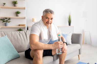 Spor salonundaki spor antrenmanından sonra tatlı su içen sporcu koltukta oturup kameraya gülümsüyor.