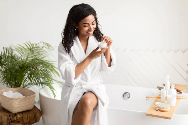 Черная леди держит крем для увлажнения сидя на ванне в ванной комнате — стоковое фото