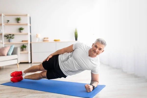 Sağlıklı yaşam tarzı kavramı. Olgun bir adam evdeki spor minderinde yan tahtada durur, oturma odasında egzersiz yapar. — Stok fotoğraf