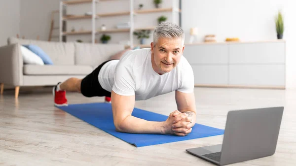 Sabah antrenmanı. Sportif orta yaşlı adam yoga tahtası yapıyor, dizüstü bilgisayarda online ders izliyor, oturma odasında egzersiz yapıyor. — Stok fotoğraf