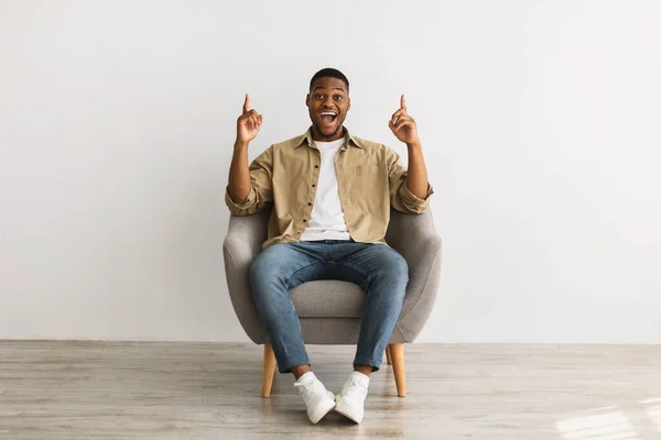 Emocionado hombre milenario africano señalando con los dedos hacia arriba sentado, fondo gris —  Fotos de Stock