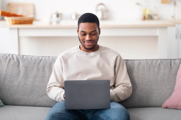 Sonriente chico negro usando el ordenador portátil, sentado en el sofá —  Fotos de Stock