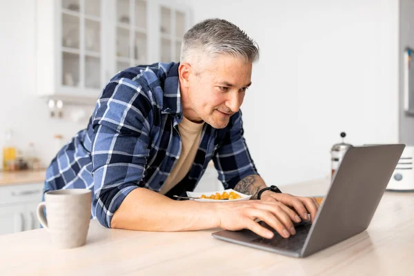 Happy starý muž relaxaci v kuchyni s notebookem a snídaní, prohlížení sociálních médií nebo čtení zpráv on-line na počítači. — Stock fotografie