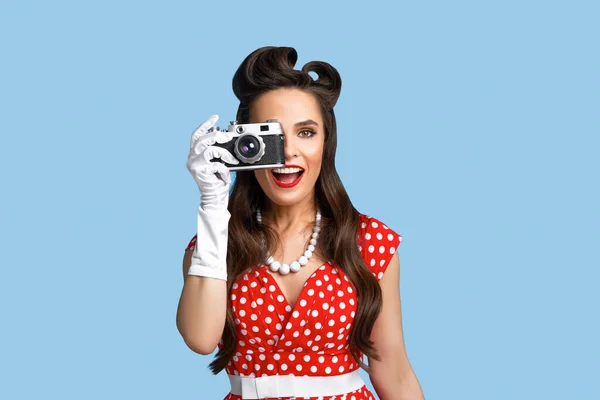 Porträtt av glada pinup kvinna i polka dot klänning ta bilder med retro kamera på blå studio bakgrund — Stockfoto