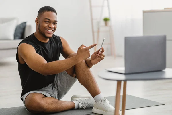 Chico negro de la aptitud que muestra Smartphone al ordenador portátil que recomienda la aplicación en interiores —  Fotos de Stock