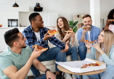 Rahatlamış çok ırklı arkadaşlar, evde parti veriyorlar, oturma odasında pizza yiyorlar, sohbet ediyorlar, gülüyorlar, birlikte vakit geçiriyorlar.