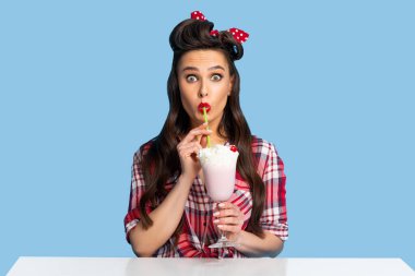 Şok olmuş esmer kadın portresi retro kıyafetli, mavi stüdyo arka planında milkshake içen.