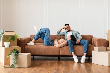 Family relaxing on couch in new home with cardboard boxes clipart