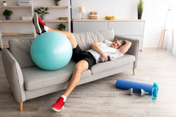 Усталость зрелый человек отдыхает на диване после активной тяжелой тренировки. Уставший спортсмен с гантелями и мячом на диване — стоковое фото