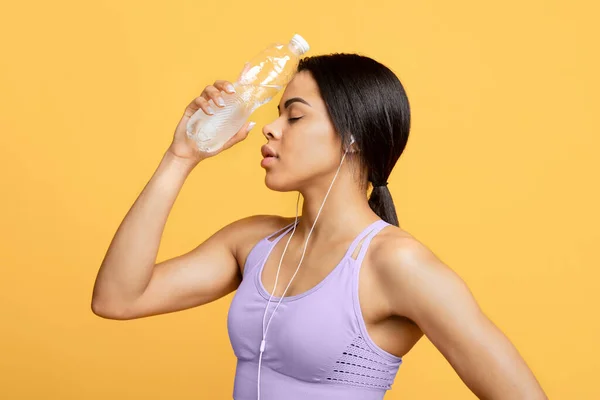 Mujer deportiva afroamericana cansada con los ojos cerrados sosteniendo botella de agua en la frente, fondo amarillo —  Fotos de Stock