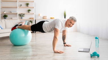 Ev içi internet eğitimi. Olgun bir adam spor topunda tahtada durur, spor videoları izler ve çalışır.