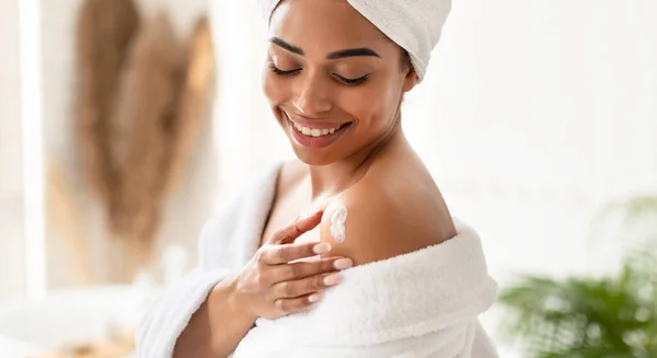 Μαύρη νεαρή γυναίκα εφαρμογή ενυδατική κρέμα στους ώμους στο μπάνιο — Φωτογραφία Αρχείου