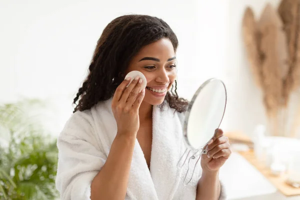 Черная женщина с помощью хлопчатобумажной панели смотрит на зеркало в ванной комнате — стоковое фото