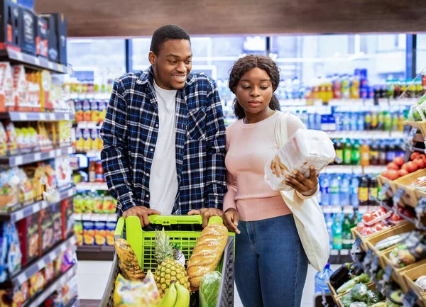 Porträtt av glada svarta par med kundvagn full av produkter som väljer mat tillsammans på moderna snabbköp — Stockfoto