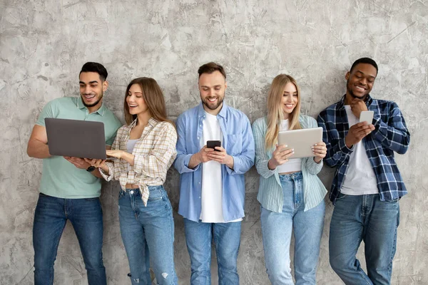 Grupo de diversos jóvenes amigos con diferentes aparatos que estudian o trabajan juntos contra la pared gris del estudio — Foto de Stock