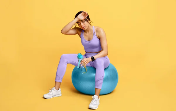 Pihenj és pihenj. Sportos fáradt fekete nő ül fitball üveg vízzel edzés után, sárga háttér — Stock Fotó