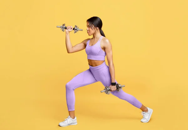 Fitness mujer afroamericana entrenando con dos mancuernas, haciendo ejercicios de embestidas para el músculo de la pierna sobre fondo amarillo —  Fotos de Stock