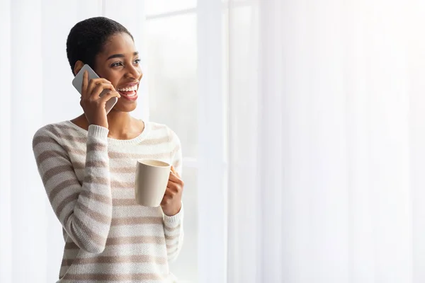 Бодрая черная женщина пьет кофе и разговаривает по телефону у окна — стоковое фото