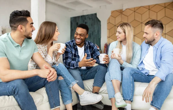 Group of cheerful multiethnic friends having party at home, sitting on couch, drinking coffee — Stock Photo, Image