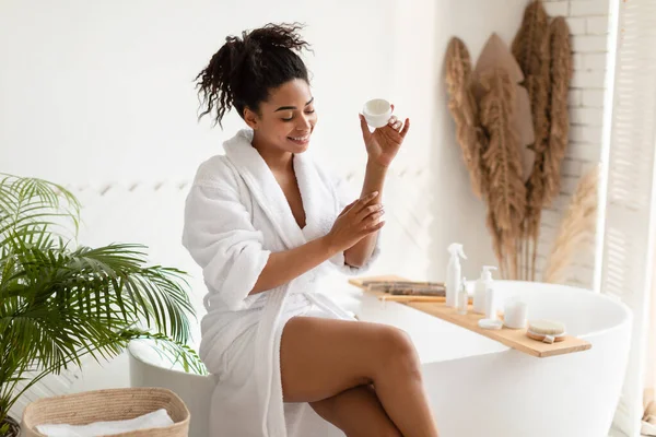 Чорна леді зволожуючі лікті застосовуючи зволожувач крем сидить у ванній кімнаті — стокове фото