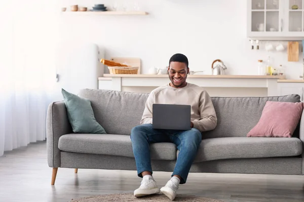 Sorrindo cara preto usando laptop, sentado no sofá — Fotografia de Stock