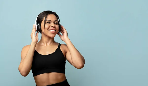 Tempo livre ativo. Senhora preta desportiva em uniforme esportivo com fones de ouvido exercitando ou dançando, fundo azul, espaço livre — Fotografia de Stock