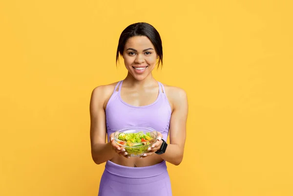 Aufgeregt afrikanisch-amerikanische fitte Frau hält Schüssel mit frischem Gemüsesalat auf gelbem Studiohintergrund — Stockfoto