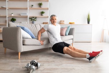 Formda kalmak için evde eğitim. Mutlu, olgun bir adam kanepeye uzanıp egzersiz yapıyor, oturma odasında çalışıyor.