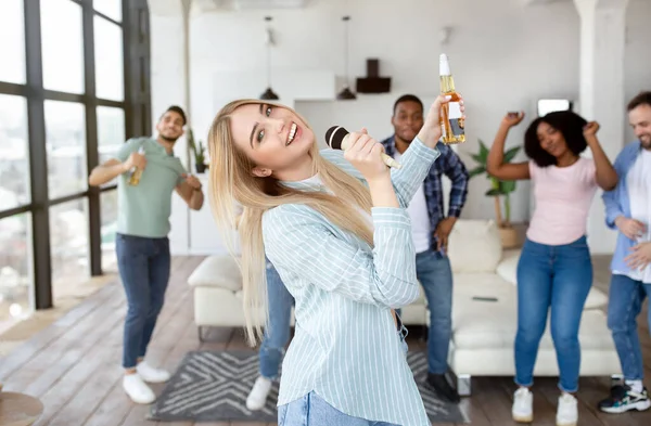 Positieve millennial blonde dame met fles bier zingen karaoke terwijl het hebben van feest met haar vrienden thuis — Stockfoto