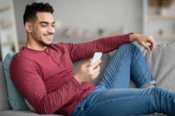 Leende arabiska killen liggande på soffan, med hjälp av smartphone — Stockfoto