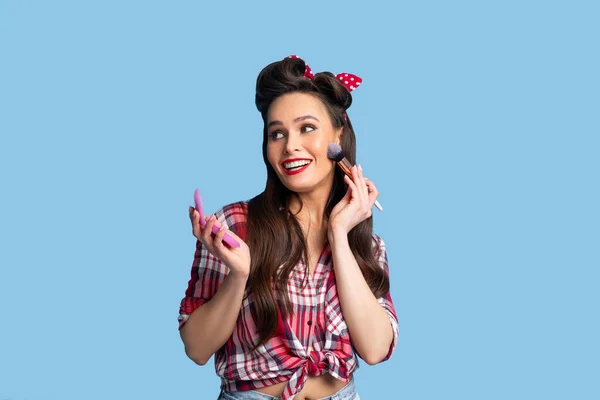 Helles Make-up. Sexy junge Pinup-Dame im Retro-Outfit setzt dekorative Kosmetik mit Pinsel über blauem Hintergrund auf — Stockfoto